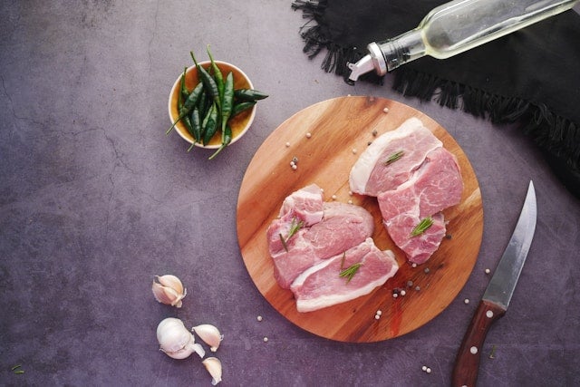 丸い木のまな板の上の二枚の豚肉と、香辛料や調理器具