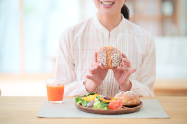 パンやサラダ、野菜ジュースの食事を摂る白いブラウス姿の女性